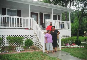 Three people stand on the steps of a white house with a porch and railing. Flowers and bushes line the garden in front, and trees are visible in the background.
