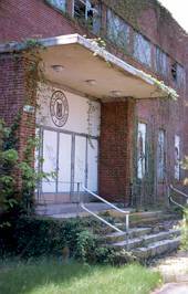 An abandoned brick building with overgrown ivy, cracked steps, and boarded-up windows; a faded emblem is visible above the entrance doors.