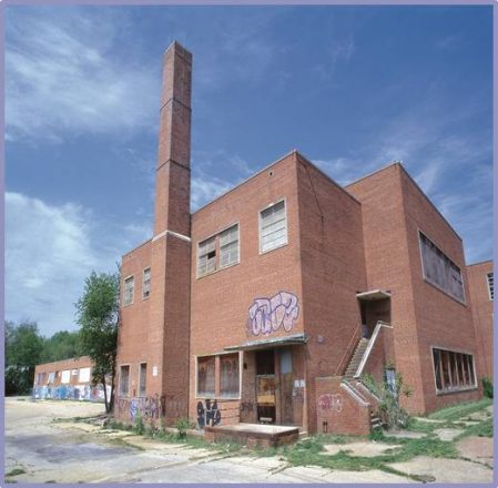 A large, abandoned red-brick building with boarded-up windows, graffiti on the walls, and a tall brick chimney, under a blue sky with scattered clouds. Overgrown vegetation surrounds parts of the building.