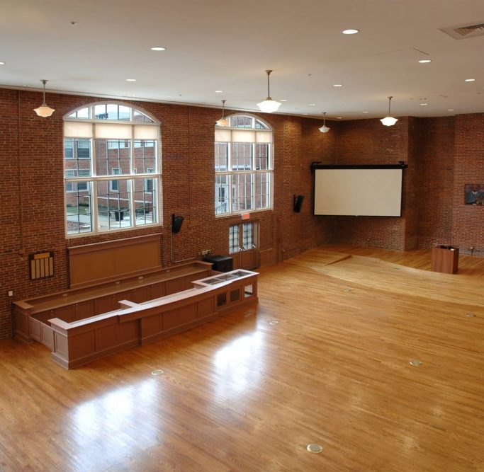 Spacious, empty hall with polished wooden floors, brick walls, large arched windows, a stage with a lectern, a projection screen, and a wooden counter area. Natural light fills the room.