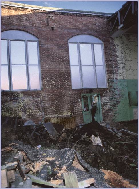 A person stands in a doorway of a damaged brick building with large windows. Debris and broken materials cover the ground, and the area appears to be in disrepair or partially destroyed.