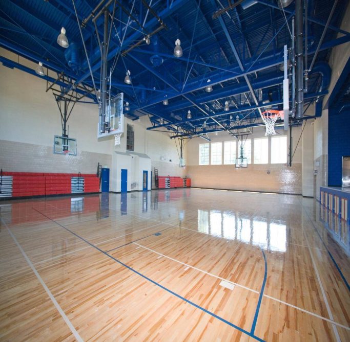 A spacious empty indoor basketball gym with shiny wooden floors, multiple basketball hoops, high blue ceiling with exposed beams, and red bleachers along the wall. Large windows let in natural light.