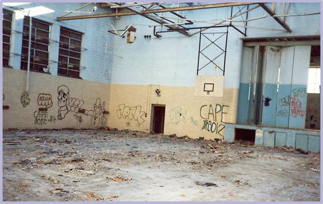 An abandoned gymnasium with broken windows, graffiti on the walls, a damaged basketball hoop, debris covering the floor, and exposed ceiling structures.