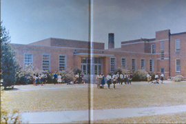 A group of people, including children and adults, gather outside a brick school building with large windows on a sunny day. The building is surrounded by shrubs and a grassy lawn.