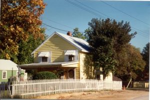 A small yellow house with white trim and striped awnings sits behind a white picket fence, surrounded by trees under a clear blue sky. The porch is partially shaded and the street is unpaved.