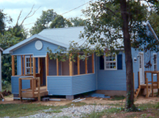 A small blue house with white trim and a covered porch is surrounded by trees. The yard has some bare earth and gravel, and the house appears to be under construction or renovation.