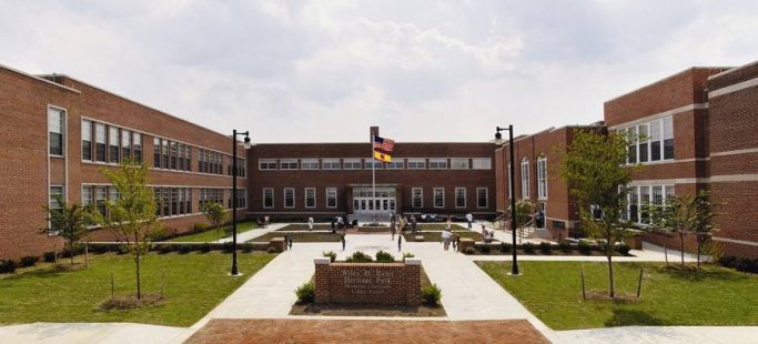A wide view of a red-brick school building with two wings and a central entrance. A courtyard with paths, grass, and a flagpole displaying the U.S. and Maryland flags is in front. Students are scattered around the courtyard.
