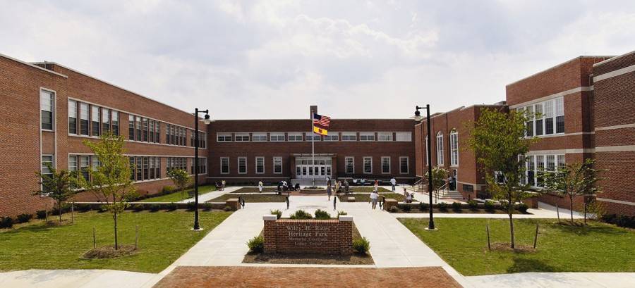 A wide view of a red-brick school building with two wings and a central entrance. A courtyard with paths, grass, and a flagpole displaying the U.S. and Maryland flags is in front. Students are scattered around the courtyard.