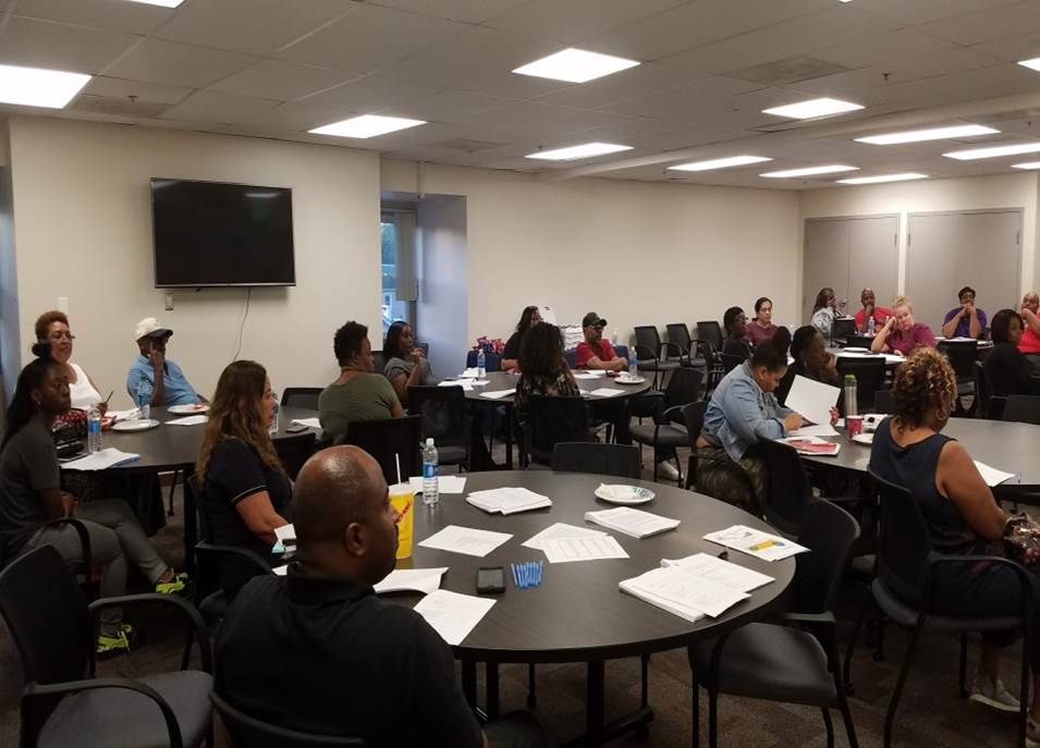 People sit in groups at round tables in a conference room, listening and looking toward the front. Papers, drinks, and snacks are on the tables, and a TV is mounted on the wall. The room is brightly lit and spacious.
