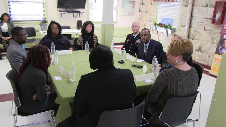 A group of nine adults sit around a rectangular table covered with a green cloth, engaged in discussion. Bottled water and name cards are on the table. The setting appears to be a community or meeting room.