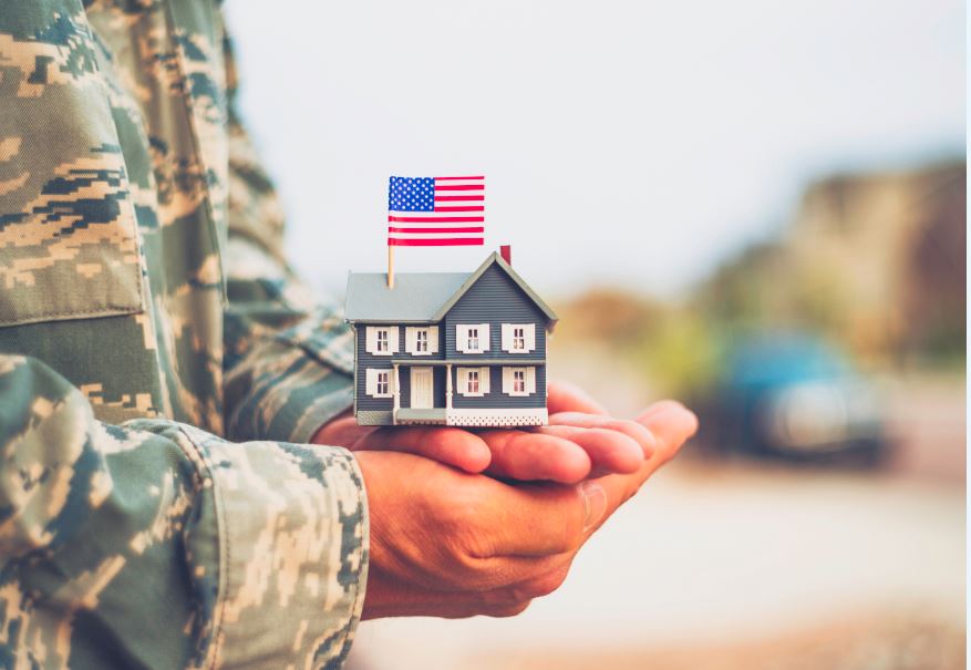 A person in military uniform holds a small model house with an American flag on top, symbolizing support for veterans, military families, or homeownership for service members.