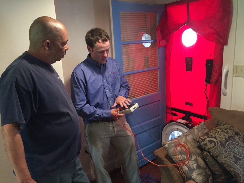 Two men stand inside a home. One holds a device connected to wires, explaining its use to the other. A red air-sealing door test setup is installed on the door, indicating an energy efficiency or air leak test.
