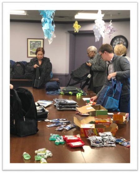 Several people stand around a table filled with snacks, toiletries, and supplies, packing items into backpacks in an indoor setting with paper decorations hanging from the ceiling.