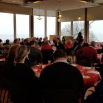 A large group of people sits around round tables covered with red tablecloths in a banquet hall, attentively listening to a speaker at the front. Large windows reveal a foggy landscape outside.