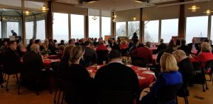 A large group of people sits around round tables covered with red tablecloths in a banquet hall, attentively listening to a speaker at the front. Large windows reveal a foggy landscape outside.