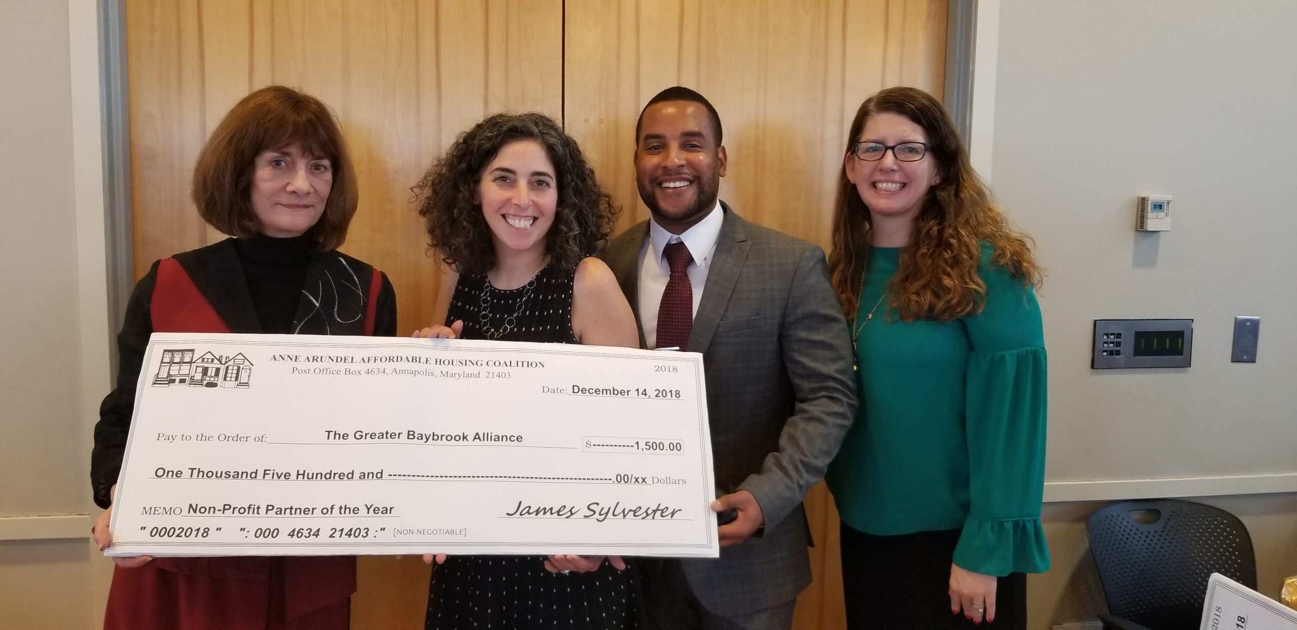 Four people stand smiling, holding a large ceremonial check made out to The Greater Baybrook Alliance for $1,500 from the Anne Arundel Affordable Housing Coalition. The photo appears to be taken at an indoor event.