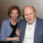 A smiling woman with curly hair and a checkered jacket sits next to a smiling man with a beard and a blue blazer, posing together against a textured background.