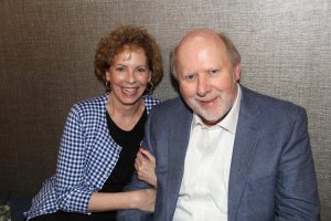 A smiling woman with curly hair and a checkered jacket sits next to a smiling man with a beard and a blue blazer, posing together against a textured background.
