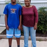 A teenage boy in a blue T-shirt and shorts stands next to a woman in a maroon top and jeans. They are smiling outdoors on a sidewalk with green bushes and a building in the background.