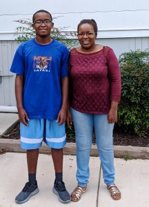 A teenage boy in a blue T-shirt and shorts stands next to a woman in a maroon top and jeans. They are smiling outdoors on a sidewalk with green bushes and a building in the background.