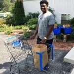 A young man smiles while holding a shopping cart with a brown paper bag in it. He is outdoors near a brick walkway, greenery, and blue plastic bins.