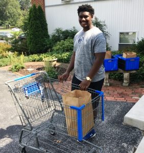A young man smiles while holding a shopping cart with a brown paper bag in it. He is outdoors near a brick walkway, greenery, and blue plastic bins.