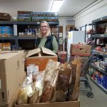 A woman stands and smiles in a food pantry, surrounded by shelves stocked with canned goods and boxes. In front of her are large boxes filled with various breads and groceries. A shopping cart is visible to the side.
