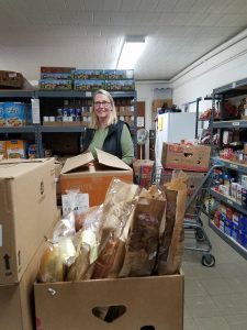 A woman stands and smiles in a food pantry, surrounded by shelves stocked with canned goods and boxes. In front of her are large boxes filled with various breads and groceries. A shopping cart is visible to the side.