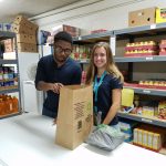 Two people stand at a table in a food pantry, packing groceries into a paper bag. Shelves behind them are stocked with canned goods, drinks, and food items. Both are smiling and appear to be volunteering.