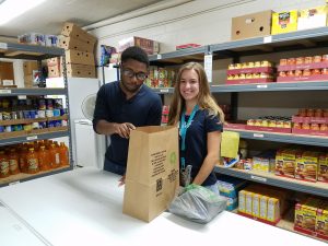 Two people stand at a table in a food pantry, packing groceries into a paper bag. Shelves behind them are stocked with canned goods, drinks, and food items. Both are smiling and appear to be volunteering.