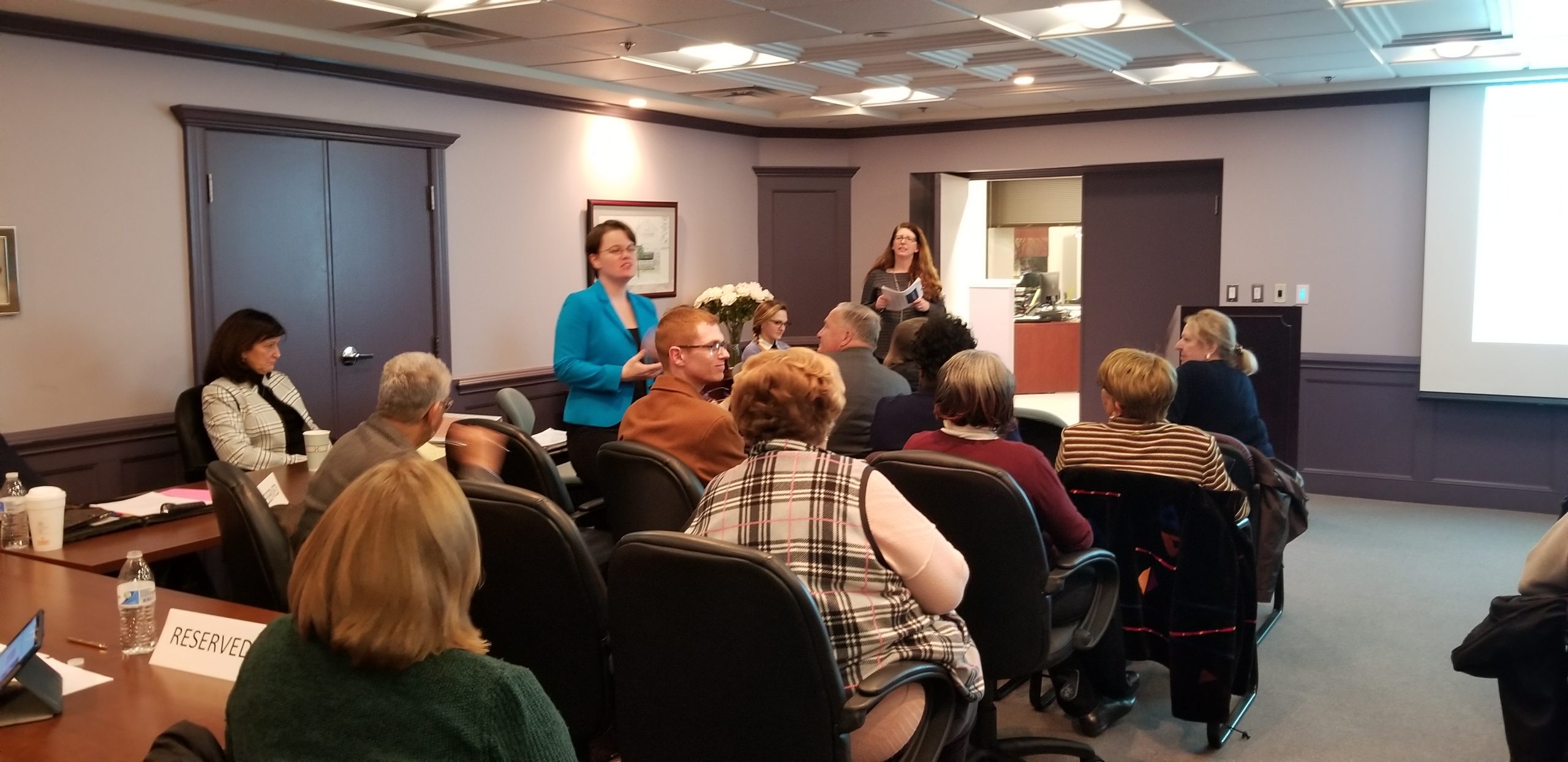 A group of people sit around a conference table in a meeting room while one woman stands, speaking, and another woman stands near a door holding papers. A "RESERVED" sign is visible on a table in the foreground.
