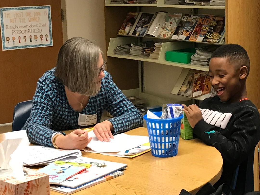A woman with gray hair and glasses sits at a table with a young boy, both smiling as they work on papers together. The table has books, papers, and a blue basket. Shelves with magazines are in the background.