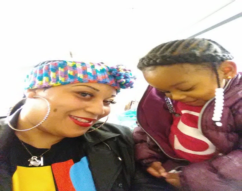 A woman wearing a colorful headband and large hoop earrings smiles at the camera beside a young girl with braided hair and beads, who is looking down and wearing a purple jacket.