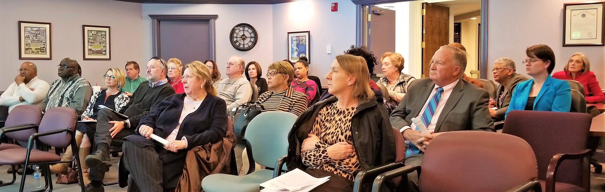 A group of adults sits attentively in a conference room, facing forward. They appear to be listening to a speaker or presentation. The room has framed pictures on the wall and a mix of colorful chairs.