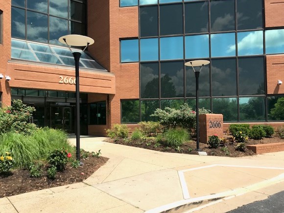 The entrance to a modern brick office building with large glass windows, the number 2666 above the door and on a sign, landscaped bushes, flowers, and two black street lamps along the walkway.
