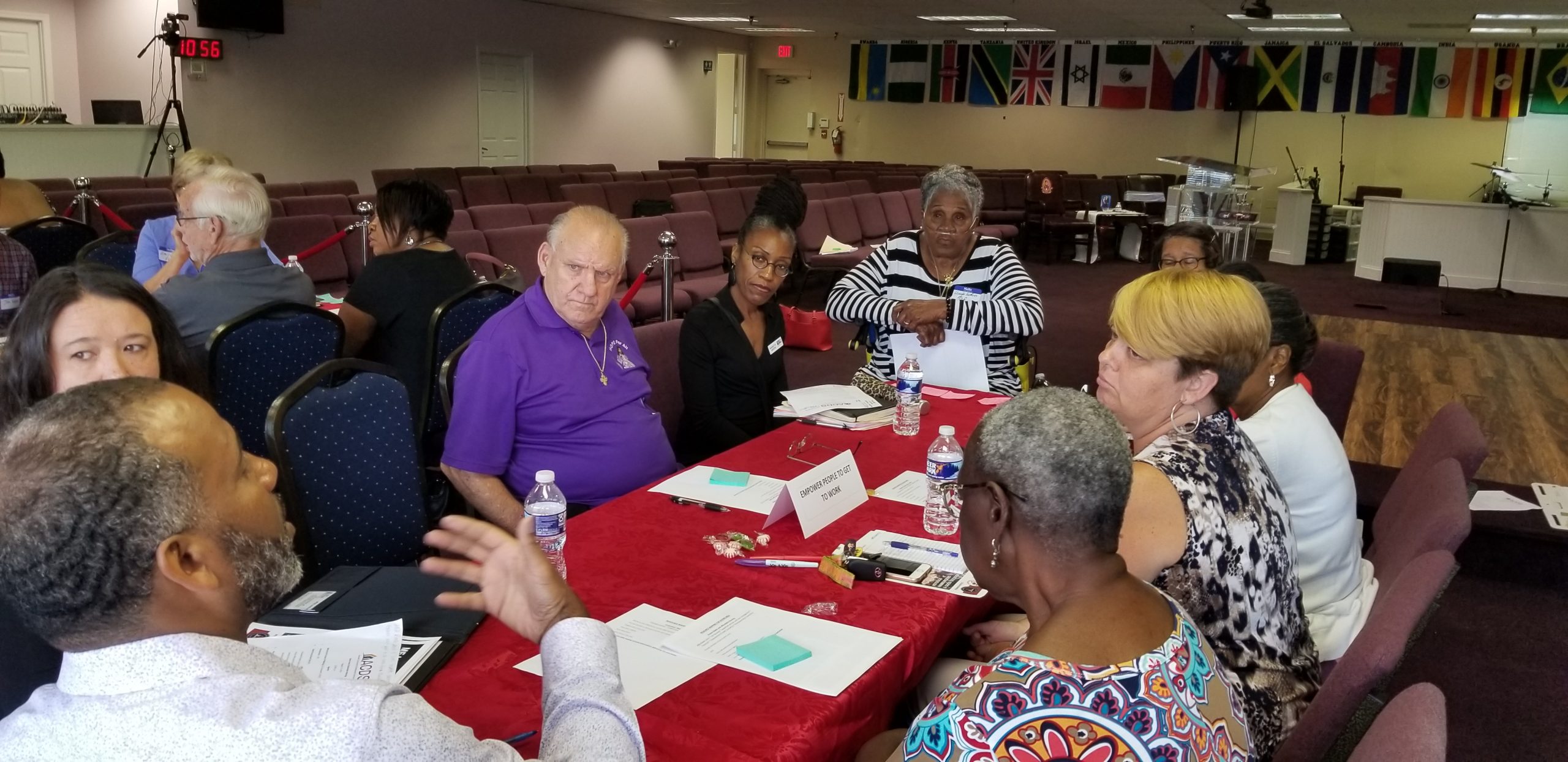 A diverse group of people sits around a red-covered table in a meeting room, engaged in discussion. Papers, water bottles, and name cards are on the table. International flags hang on the wall in the background.