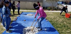 A group of children stands on grass, some watching while one girl uses tongs to move shells on a blue tarp. Other kids wait nearby, and a woman stands in the background next to an orange basket.