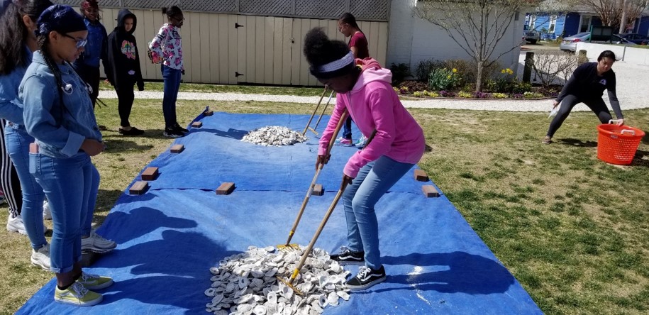 A group of children stands on grass, some watching while one girl uses tongs to move shells on a blue tarp. Other kids wait nearby, and a woman stands in the background next to an orange basket.