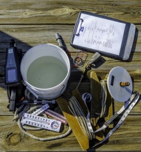 A white bucket of water, thermometer, measuring tapes, ropes, wooden boards, a meter, and a clipboard with papers are arranged on a wooden dock in bright sunlight.