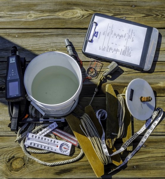 A white bucket of water, thermometer, measuring tapes, ropes, wooden boards, a meter, and a clipboard with papers are arranged on a wooden dock in bright sunlight.
