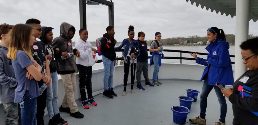 A group of students stands in a line on a boat deck, listening to an instructor in a blue jacket. Two blue buckets are on the floor, and another adult stands nearby holding a phone. Water and shoreline are visible in the background.