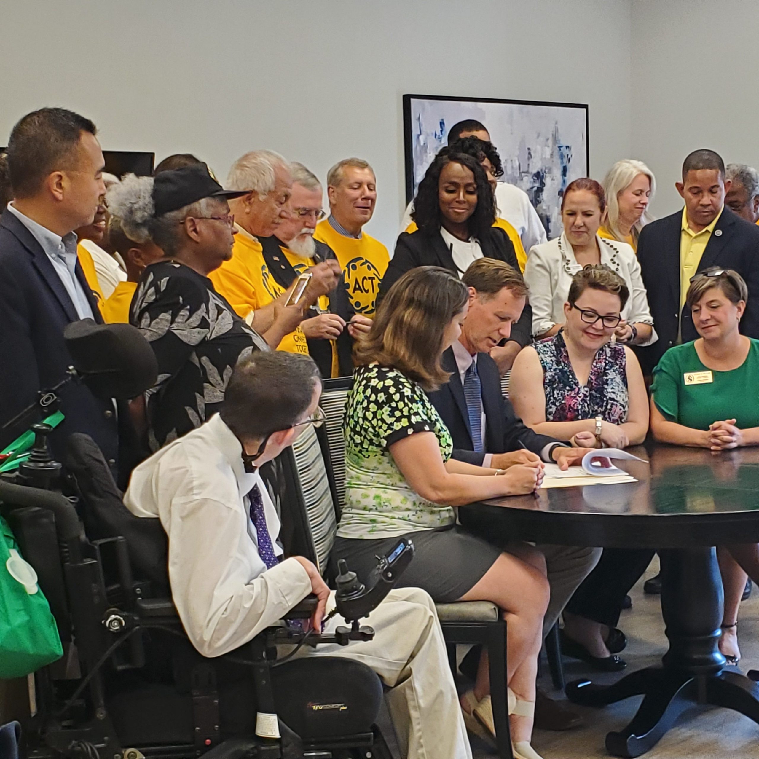 A group of people, including advocates and officials, gather around a table as two seated individuals sign documents. One person in a wheelchair is in the foreground. Others stand behind, some wearing yellow shirts, smiling and observing.