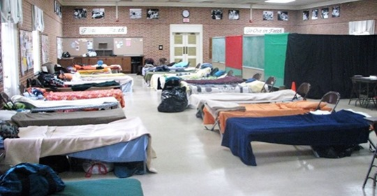 A large room with many cots lined up, each with blankets and personal items. The room has a tiled floor, brick walls, and a banner reading “Go Out in Faith.” It appears to be a temporary shelter or sleeping area.