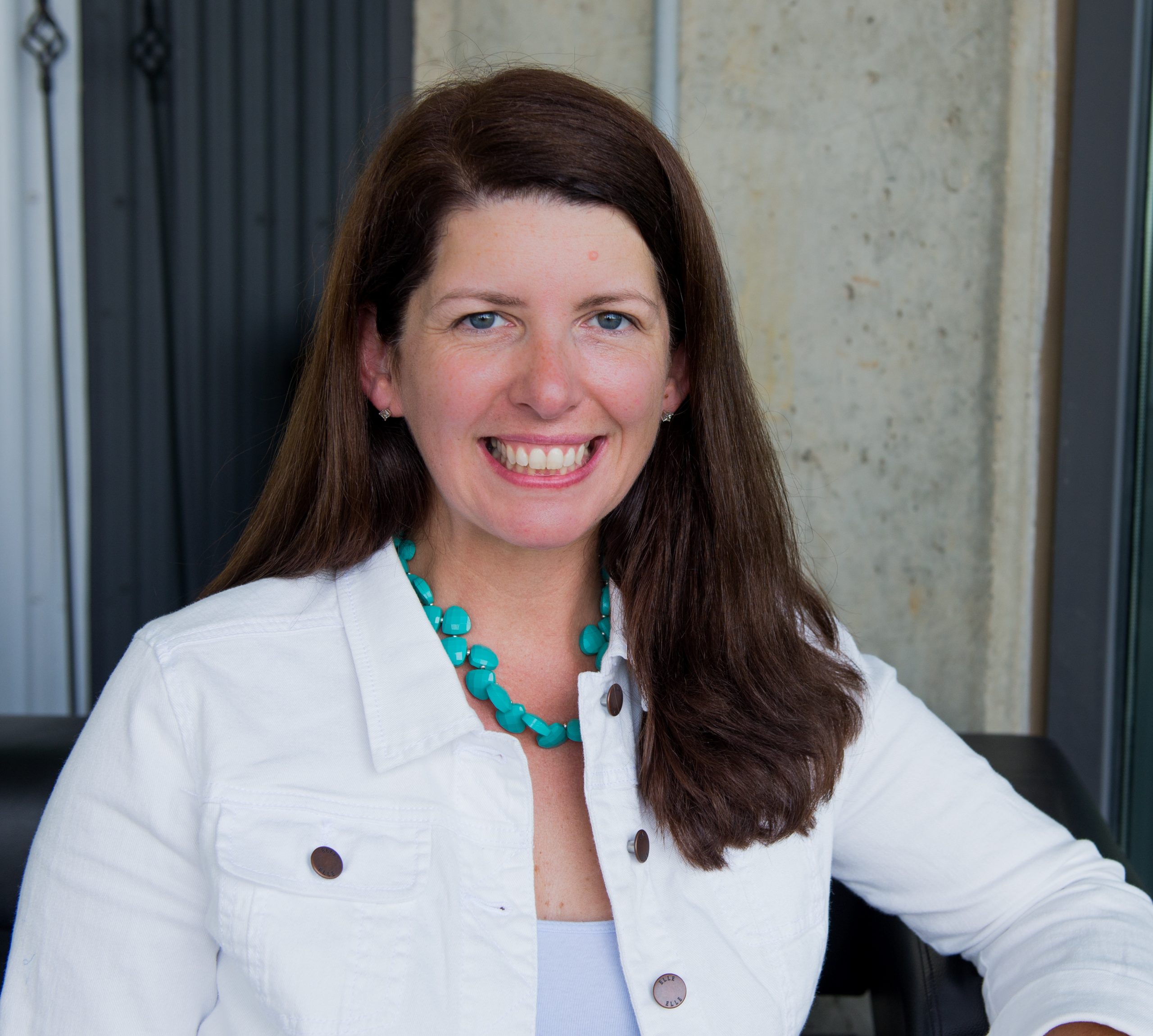 A woman with long brown hair, wearing a white jacket and turquoise necklace, sits and smiles at the camera. The background includes a concrete wall and a dark door.