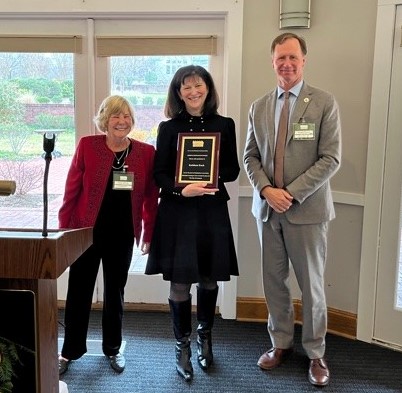 Three adults stand indoors near windows. The woman in the center holds a plaque and smiles. The woman on the left wears a red jacket, and the man on the right wears a gray suit. All are smiling, appearing at an award event.