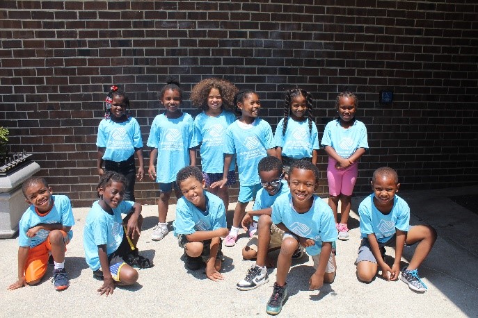 A group of eleven smiling children wearing matching light blue shirts pose together outside in front of a brick wall, some standing and some kneeling on the ground.