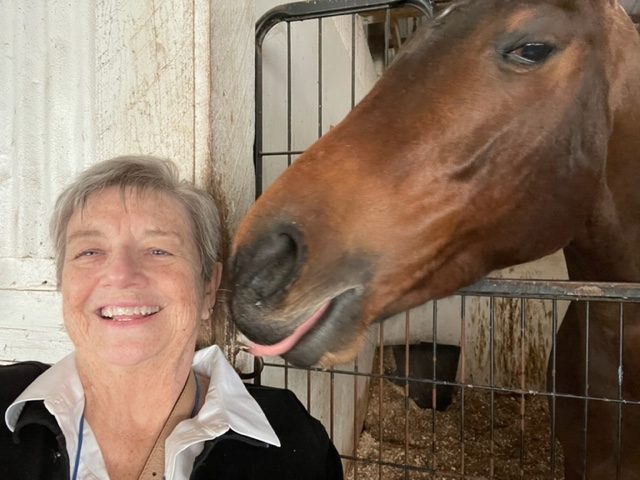 A smiling older woman stands in front of a stable, while a brown horse behind her playfully reaches its head over a gate, appearing to lick her cheek.