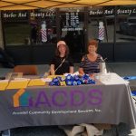 Two women sit at an outdoor booth for Arundel Community Development Services, Inc., with snacks and papers on the table, under a tent, in front of a barber and beauty shop.