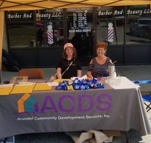 Two women sit at an outdoor booth for Arundel Community Development Services, Inc., with snacks and papers on the table, under a tent, in front of a barber and beauty shop.