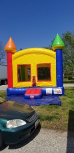 A colorful inflatable bounce house with red, yellow, green, and blue sections sits on grass next to a parked green car under a clear blue sky.